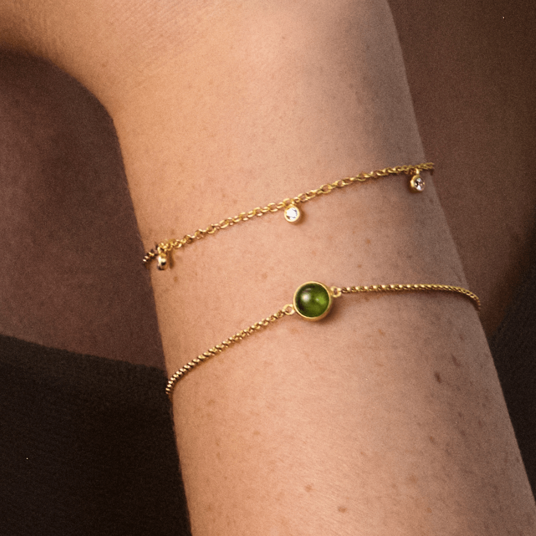 Primini armbånd med grønn cabochon-krystall båret på håndleddet, lagdelt med et tynt sølvarmbånd med to klare steiner
