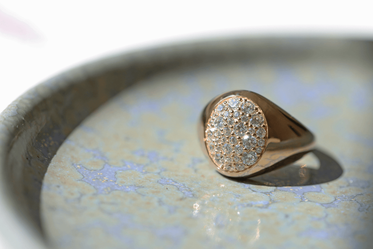 Multipave signetring fra Julie Sandlau i gull, nærbilde i keramisk skål med diamantfelt i fokus.