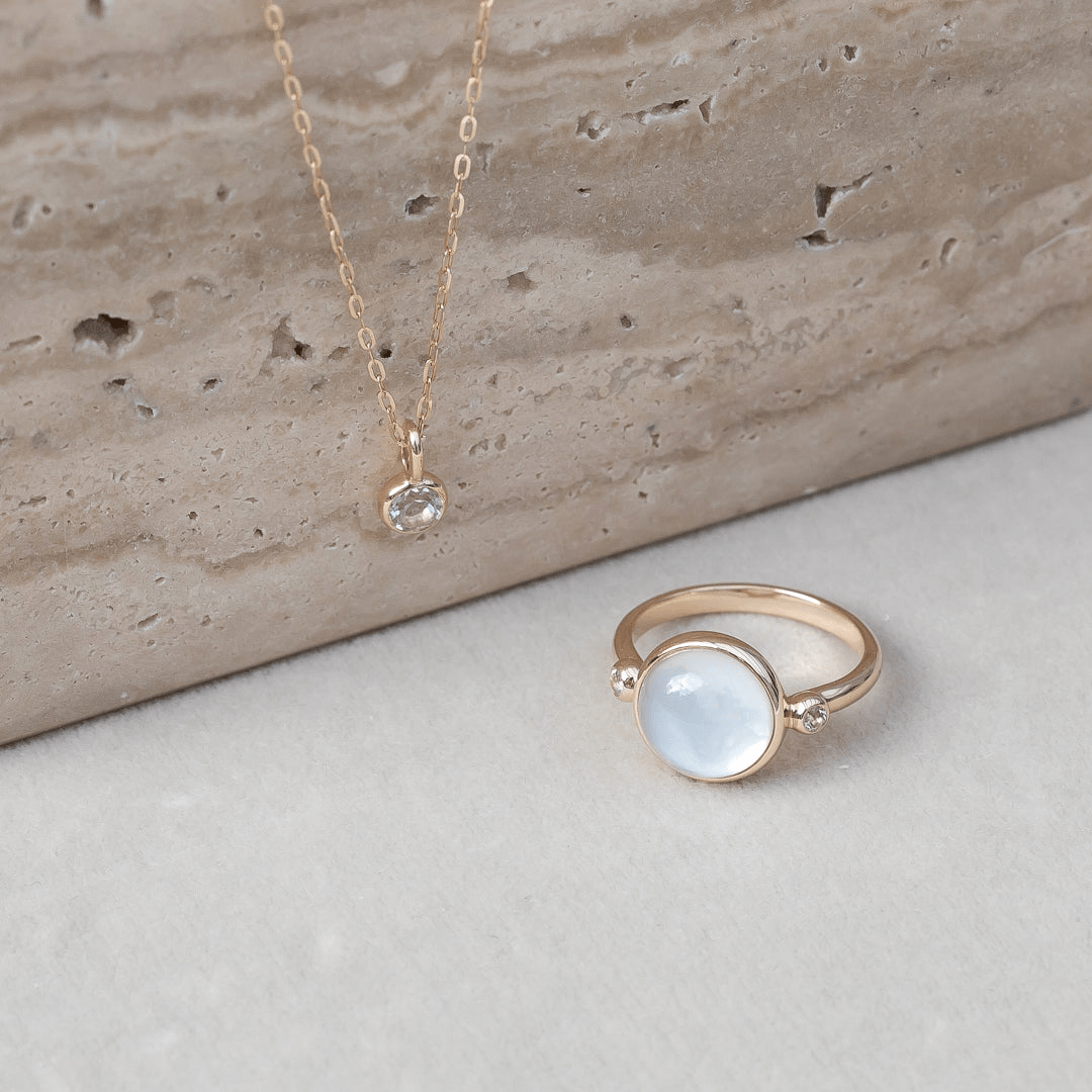 Carla Ring i gull med hvit cabochon-slipt stein, foto ved siden av en gullkjede med anheng