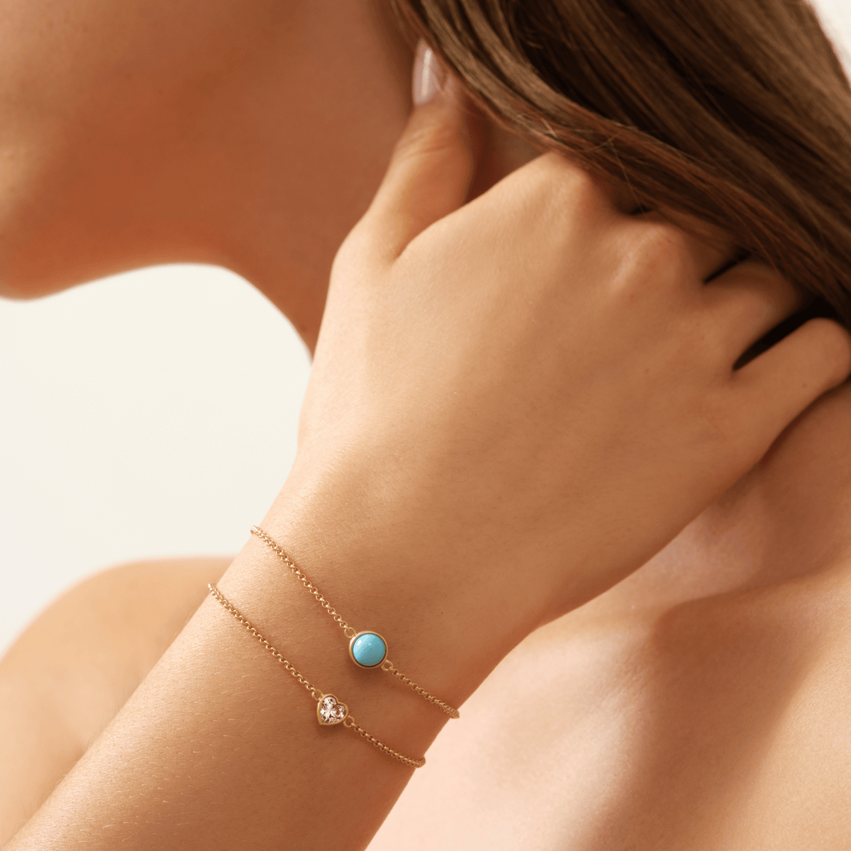 Primini armbånd med turkisblå cabochon-krystall båret på håndleddet, lagdelt med et armbånd med hjerteformet klar stein