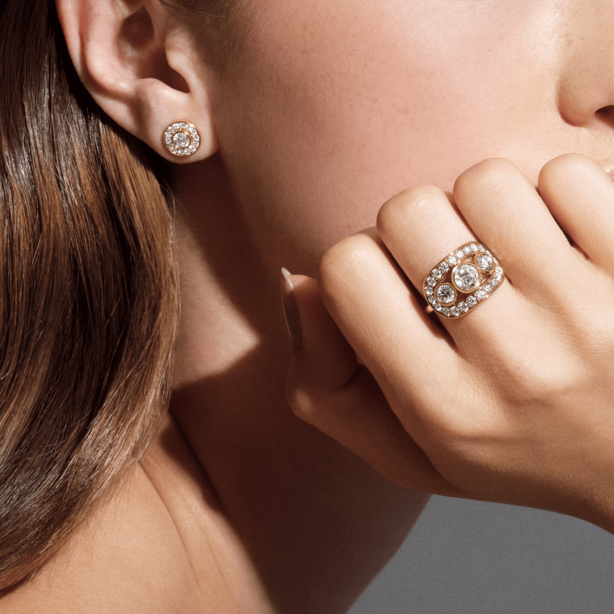 Heritage Trio Ring båret på hånden holdt opp mot øret, med en diamantbesatt stud-ørering synlig i bakgrunnen.