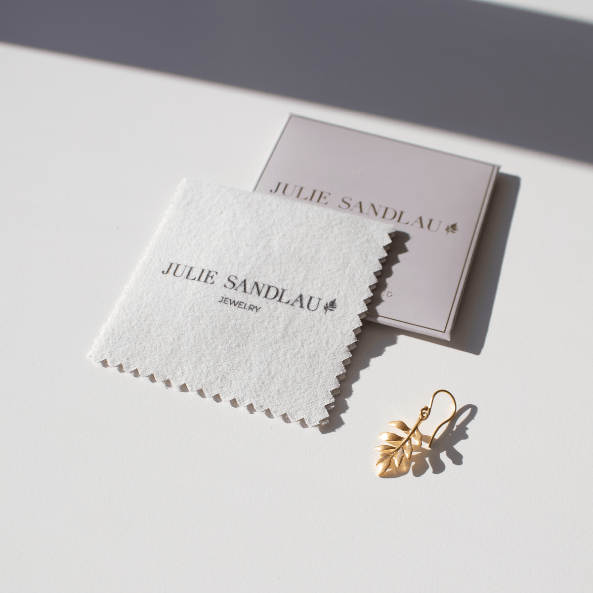 Julie Sandlau pusseklut med logo og eske ved siden av en gullfarget øredobb, produktbilde